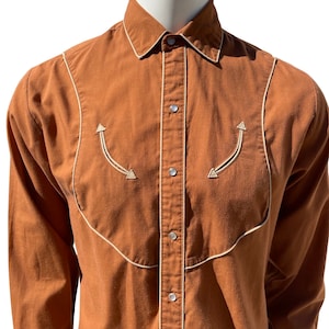 Puede incluir: Una camisa marrón de manga larga con costuras blancas alrededor del cuello, los bolsillos y el canesú. La camisa tiene un cierre de botones en la parte delantera y un cuello puntiagudo.