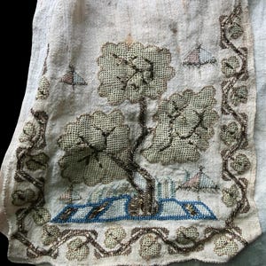 May include: Close-up of a vintage embroidered textile fragment. The design features a tree with a textured canopy, a blue and white structure, and small, stylized birds. The border is adorned with a vine-like pattern. The color palette includes beige, brown, and blue.