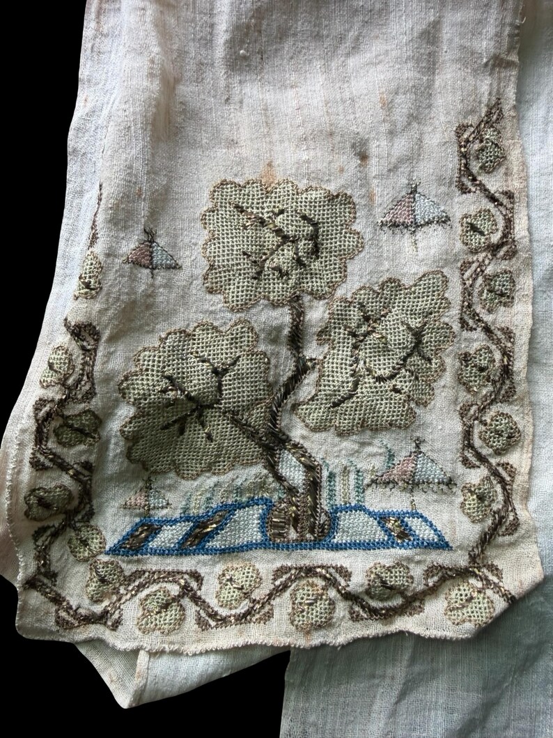 May include: Close-up of a vintage embroidered textile fragment. The design features a tree with a textured canopy, a blue and white structure, and small, stylized birds. The border is adorned with a vine-like pattern. The color palette includes beige, brown, and blue.