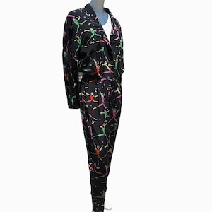 May include: Black jumpsuit with a colorful abstract print featuring figures and lines. The jumpsuit has a long sleeve, a collared neckline, and a button closure.