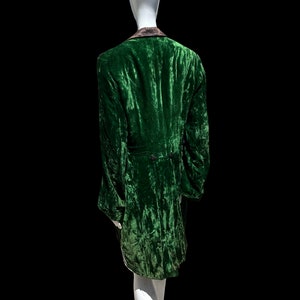 K&ouml;nnte beinhalten: Eine gr&uuml;ne Samtjacke mit schwarzem Kragen und einem langen, flie&szlig;enden Design. Die Jacke befindet sich auf einem wei&szlig;en Mannequin.