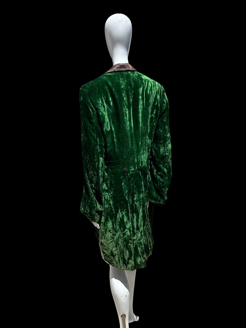 K&ouml;nnte beinhalten: Eine gr&uuml;ne Samtjacke mit schwarzem Kragen und einem langen, flie&szlig;enden Design. Die Jacke befindet sich auf einem wei&szlig;en Mannequin.