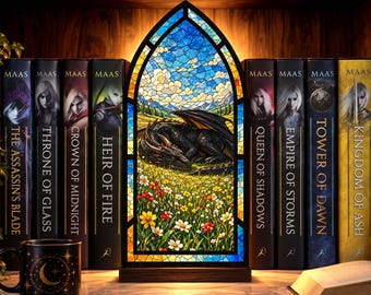 May include: A stained glass lamp shaped like a pointed arch window, featuring a black dragon sleeping in a field of colorful flowers under a blue sky. It is flanked by a row of books by Maas, including Throne of Glass and Kingdom of Ash.