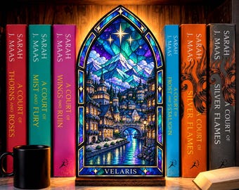 May include: An arched, stained-glass style lamp featuring a colorful illustration of a city with a river, bridge, and mountains under a starry sky. The base of the lamp displays the text VELARIS. It is positioned between several books.