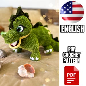 Pode incluir: Brinquedo de peluche de dinossauro verde em crochê com dentes amarelos e olhos brancos. O brinquedo está sentado em um cobertor marrom. Padrão de crochê PDF em inglês.