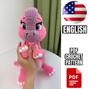 May include: Crochet pattern for a pink dinosaur amigurumi toy. The pattern is available in PDF format and is written in English. The image shows the finished toy and a hand holding it.