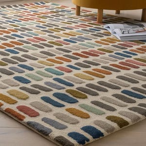 May include: A rectangular area rug with a repeating pattern of colorful, oblong shapes in shades of blue, green, yellow, orange, and gray on a cream background. The rug is placed on a light wood floor, with a yellow chair and a magazine visible.