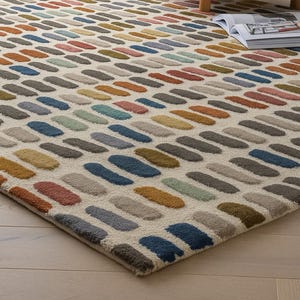 Puede incluir: Una alfombra rectangular con un fondo color crema y un patrón repetitivo de formas oblongas coloridas. Las formas son en tonos de azul, verde, naranja, gris y marrón. La alfombra está colocada sobre un suelo de madera clara.
