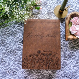 May include: A brown leather-bound Bible with the words "The Holy Bible" in script. The cover features a floral design and the name "Rebecca Kathryn Wilson." The Bible rests on a white lace surface, with flowers and a candle holder nearby.