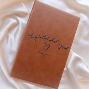 May include: A brown leather journal with the text "Cling to that which is good" and a heart symbol. The text "Romans 12:9" is below the heart. The journal is rectangular and closed.