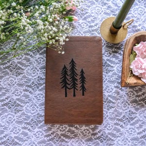 May include: A brown leather journal with a black tree design. The journal is rectangular and closed, with a simple illustration of three pine trees. The background includes white lace fabric and floral accents.