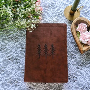 May include: A brown leather journal with three embroidered pine trees. The journal is rectangular and has a stitched border. The background includes white lace, flowers, and a candle holder.