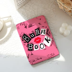 Può includere: Una custodia per telefono rosa con la scritta "Burn Book" in un design a blocchi in bianco e nero, con una grafica di labbra rosse. La custodia presenta un motivo di scarabocchi e simboli neri.