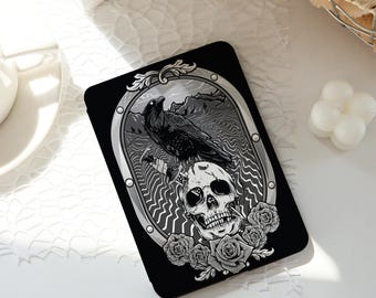 May include: Black rectangular phone case featuring a gothic design. The artwork includes a raven perched above a skull, set within an oval frame. Gray roses surround the skull, with a mountain landscape in the background.