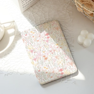 Op de afbeelding: Een rechthoekig notitieboek met een bloemenpatroon, met roze, gele en oranje bloemen op een witte achtergrond. Het notitieboek ligt op een wit oppervlak. De achtergrond is zacht en wazig.