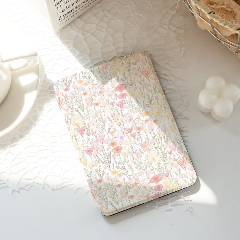 Op de afbeelding: Een rechthoekig notitieboek met een bloemenpatroon, met roze, gele en oranje bloemen op een witte achtergrond. Het notitieboek ligt op een wit oppervlak. De achtergrond is zacht en wazig.