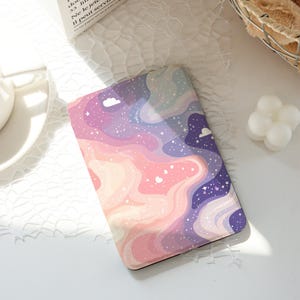 Könnte beinhalten: Ein rechteckiges Notizbuch mit einem farbenfrohen, verträumten Design. Das Cover zeigt ein wirbelndes Muster aus rosa, lila und blauen Farbtönen, mit weißen Wolken und Sternen, die über die Oberfläche verstreut sind. Das Notizbuch liegt auf einer weißen Oberfläche.