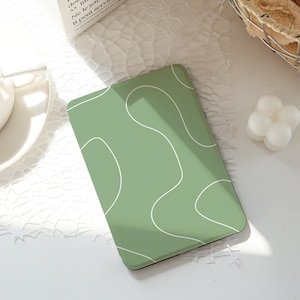 May include: A rectangular, sage green phone case with a white, abstract, wavy design. The case is on a white surface with other objects in the background.