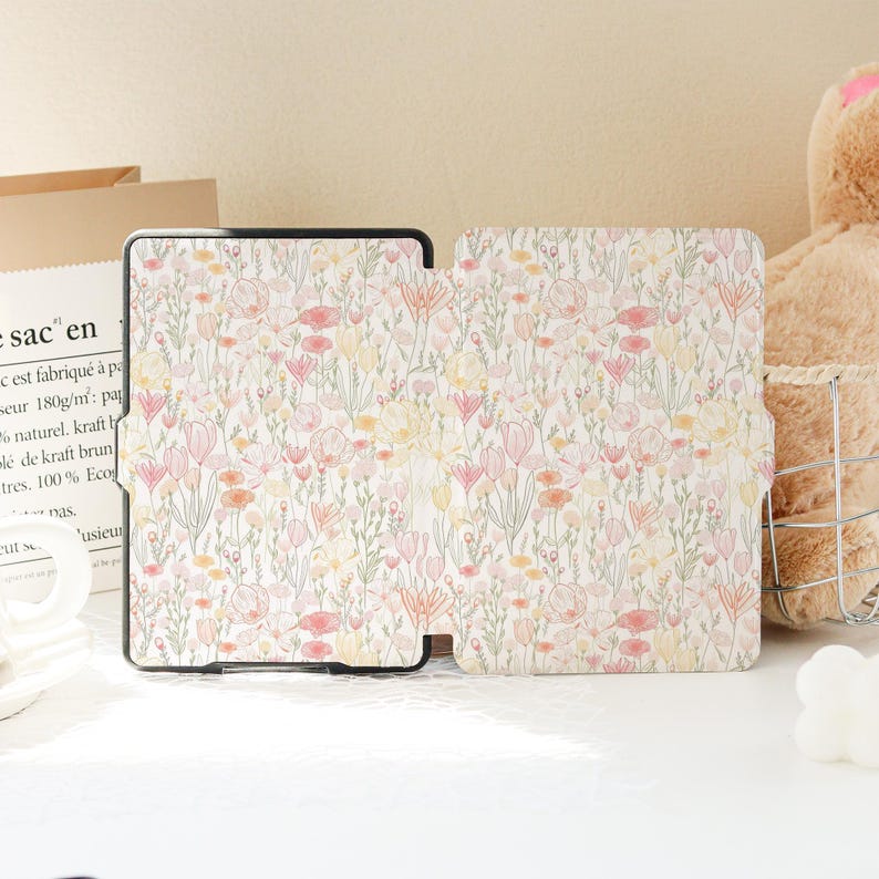 Op de afbeelding: Een tablet hoesje met een bloemenpatroon in roze, geel en oranje op een cr&egrave;mekleurige achtergrond. De hoes is open en toont de binnenkant en het bloemmotief. De hoes is zwart.