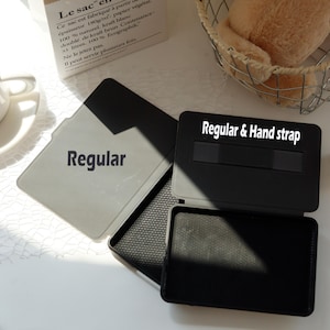 Op de afbeelding: Een open zwarte en grijze hoes met de tekst "Regular & Hand strap" bovenaan. De hoes is open en onthult een grijs interieur met het woord "Regular" op de hoes. De hoes ligt op een wit oppervlak.