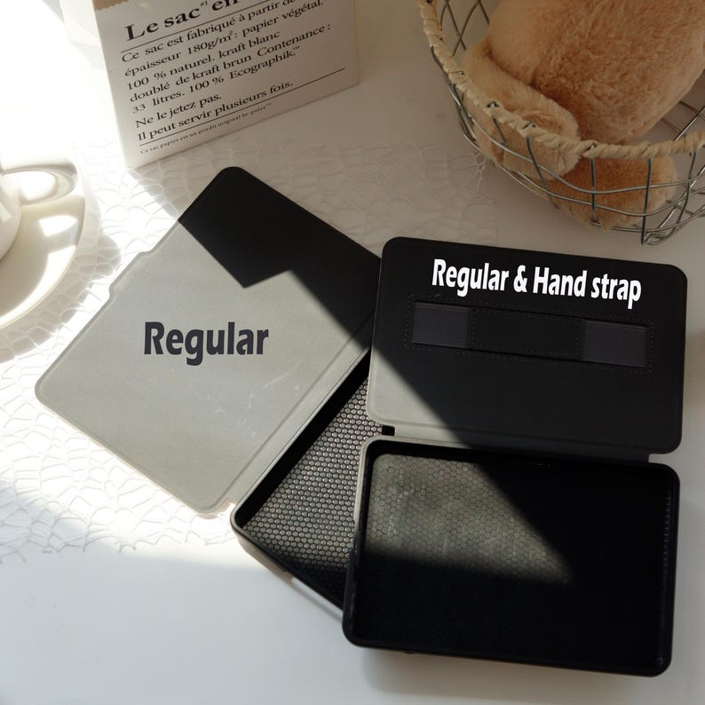 Op de afbeelding: Een open zwarte en grijze hoes met de tekst "Regular & Hand strap" bovenaan. De hoes is open en onthult een grijs interieur met het woord "Regular" op de hoes. De hoes ligt op een wit oppervlak.