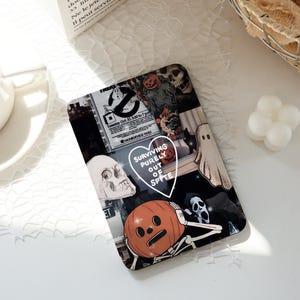 Könnte beinhalten: Eine rechteckige Handyhülle mit einer Collage zum Thema Halloween. Das Design umfasst Totenköpfe, Geister, Kürbisse und den Text "SURVIVING PURELY OUT OF SPITE". Die Hülle hat eine glänzende Oberfläche und einen schwarzen Hintergrund.