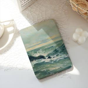 May include: A rectangular e-reader case featuring a printed oil painting of a turbulent ocean scene with crashing white waves, deep teal water, and a soft yellow and pale blue sky. The case has rounded corners and a smooth, matte finish.