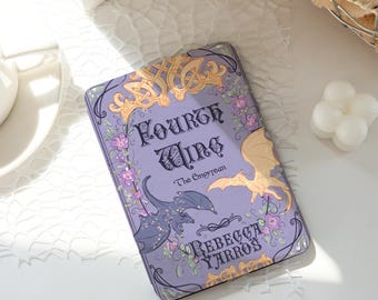 May include: A rectangular, lavender-colored book cover with the title "Fourth Wing" and author "Rebecca Yarros" in ornate lettering. The cover features illustrations of dragons, flowers, and decorative gold accents.