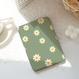 Puede incluir: Una funda de teléfono verde con un patrón repetido de margaritas crema con centros naranjas. La funda rectangular está hecha de un material liso. Las margaritas están espaciadas uniformemente.