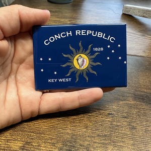 May include: A blue and yellow magnet with the text "Conch Republic" and "Key West" and a yellow sun with a conch shell inside. The text "1828" is also on the magnet.
