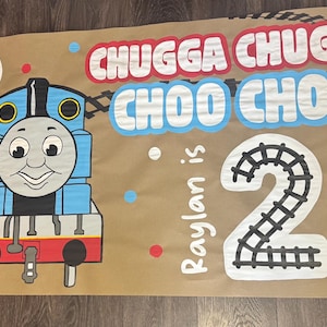 Könnte beinhalten: Braunes Banner mit einem blauen Zeichentrickzug und dem Text "Chugga Chugga Choo Choo". Die Zahl "2" und der Text "Raylan is" sind ebenfalls vorhanden. Rote, weiße und blaue Ballons sind ebenfalls abgebildet.
