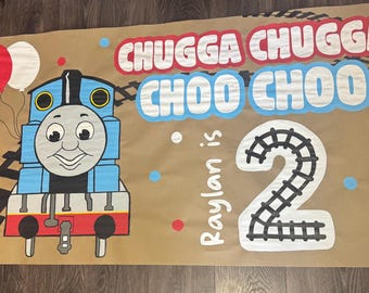 Può includere: Uno striscione marrone con un treno blu dei cartoni animati e la scritta "Chugga Chugga Choo Choo". Lo striscione include anche il numero "2" e la scritta "Raylan is". Sono presenti anche palloncini rossi, bianchi e blu.