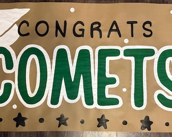May include: A brown banner with the words "CONGRATS COMETS" in black and green. A white graduation cap is on the left. Black stars are at the bottom, and white dots are scattered around the text.