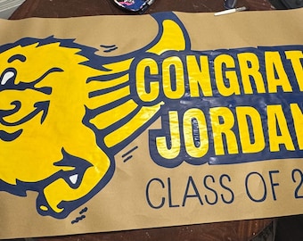 May include: A banner with a yellow cartoon mascot and the words "CONGRATS JORDAN! CLASS OF 2025" in yellow and navy blue. The banner is brown and appears to be made of paper.