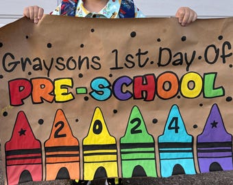 Puede incluir: Un cartel de papel marrón con el texto "Graysons 1st Day of Pre-School 2024" escrito en marcador negro. El cartel está decorado con dibujos de colores de lápices de colores.