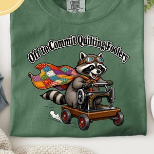 May include: A moss green t-shirt featuring a graphic of a raccoon wearing goggles and a colourful patchwork cape, riding a sewing machine on wheels. The text above the raccoon reads, Off to Commit Quilting Foolery, in a bold, black, arched font.