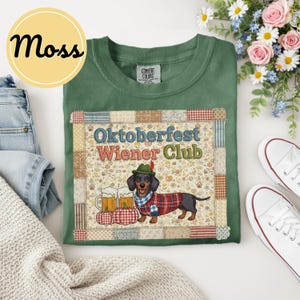 May include: A moss green t-shirt featuring a graphic of a dachshund wearing a traditional Alpine hat and a red plaid sweater. The design includes two beer mugs, pumpkins, and the text Oktoberfest Wiener Club, all framed by a patchwork border.