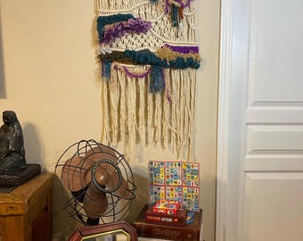 May include: A wooden table with a variety of board games and card games, including Sequence, Uno, and a vintage fan. There is also a macrame wall hanging with a floral design.