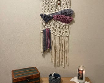 May include: A macrame wall hanging with a natural wood branch at the top. The macrame is made with cream colored yarn and features blue, purple, and pink accents. The wall hanging is displayed on a wall with a wooden shelf below it. The shelf is decorated with a wooden box, a ceramic bowl, and a candle.