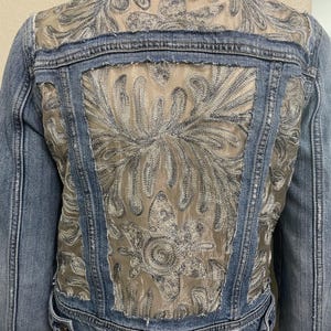 May include: Light blue denim jacket with a decorative embroidered back panel. The back features a sheer, beige fabric overlaid with silver and gray floral embroidery. The jacket has a classic collar, button closures, and long sleeves.