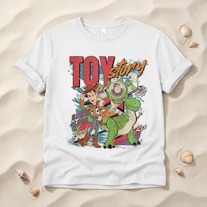 May include: A white short-sleeve t-shirt featuring a graphic print of Woody, Buzz Lightyear, and Rex from Toy Story. The design includes the text "TOY story", "SINCE 1995", "Woody", "Buzz", and "Rex" in a colorful, distressed, retro-style illustration.