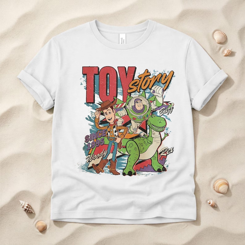 May include: A white short-sleeve t-shirt featuring a graphic print of Woody, Buzz Lightyear, and Rex from Toy Story. The design includes the text "TOY story", "SINCE 1995", "Woody", "Buzz", and "Rex" in a colorful, distressed, retro-style illustration.