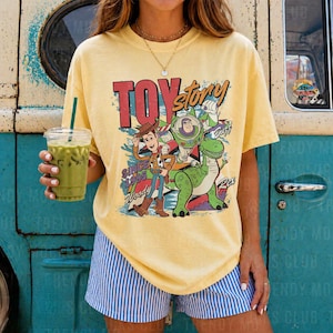 May include: A pale yellow short-sleeve t-shirt featuring a graphic print of Woody, Buzz Lightyear, and Rex from Toy Story with the text TOY story SINCE 1995. The shirt is paired with blue and white vertical striped shorts.