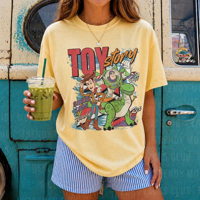 May include: A pale yellow short-sleeve t-shirt featuring a graphic print of Woody, Buzz Lightyear, and Rex from Toy Story with the text TOY story SINCE 1995. The shirt is paired with blue and white vertical striped shorts.