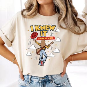 K&ouml;nnte beinhalten: Ein cremefarbenes T-Shirt mit einer Grafik einer Person mit langem Haar, die ein gelbes kariertes Hemd und eine Hose mit Kuhflecken tr&auml;gt und eine Gitarre h&auml;lt. Der Text lautet I KNEW IT und I KNEW YOU, umgeben von einem roten Hut, Noten, Sternen und Wolken.
