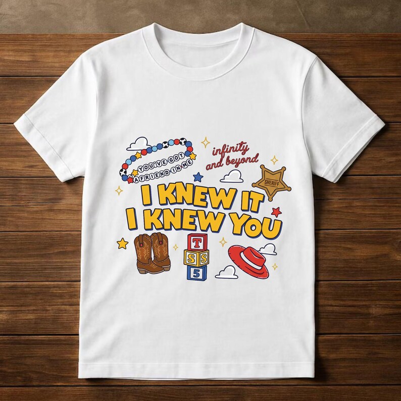 May include: A white short-sleeve t-shirt featuring a graphic print with the text I KNEW IT I KNEW YOU, infinity and beyond, and YOU'VE GOT A FRIEND IN ME. The design includes a beaded bracelet, a sheriff badge, cowboy boots, blocks, and a red hat.