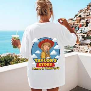K&ouml;nnte beinhalten: Ein wei&szlig;es T-Shirt mit einem Grafikdruck auf der R&uuml;ckseite. Das Design zeigt eine Zeichentrickfigur mit blonden Z&ouml;pfen und rotem Hut vor einem blauen kreisf&ouml;rmigen Hintergrund mit wei&szlig;en Wolken. Der Text lautet Taylor's Story und I knew it I knew you.