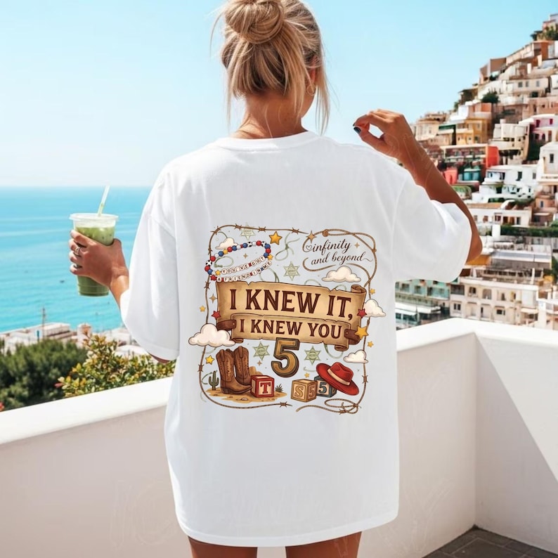 May include: A white t-shirt featuring a graphic on the back with the text "I KNEW IT, I KNEW YOU" and "infinity and beyond". The design includes a beaded bracelet, a large number 5, cowboy boots, a red hat, and alphabet blocks.