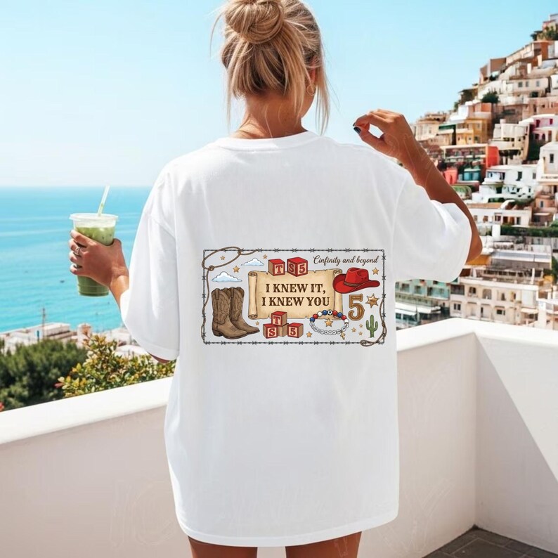以下が含まれることがあります： 背面にグラフィックがプリントされた白いTシャツ。スクロールには「I KNEW IT, I KNEW YOU」というテキストが書かれています。デザインには、カウボーイブーツ、赤い帽子、アルファベットのブロック、ビーズのブレスレット、サボテン、数字の5のイラストが含まれています。