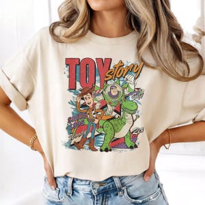 May include: A cream-colored short-sleeve t-shirt featuring a graphic print of Woody, Buzz Lightyear, and Rex from Toy Story. The design includes the text "TOY story", "SINCE 1995", "Woody", "Buzz", and "Rex" in a colorful, distressed artistic style.